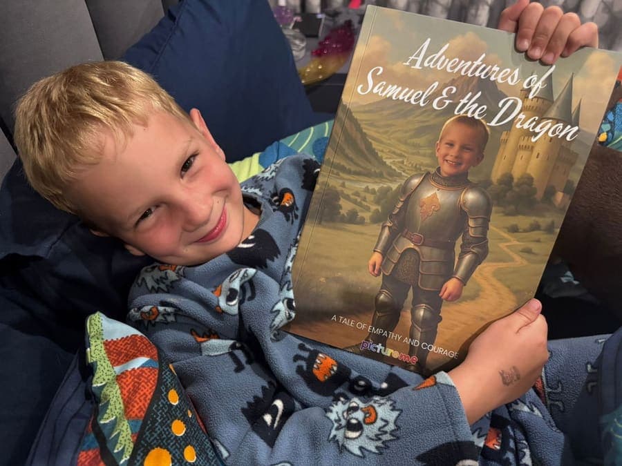 A boy snuggled in a blanket grinning next to his personalised Adventures of Samuel & the Dragon book