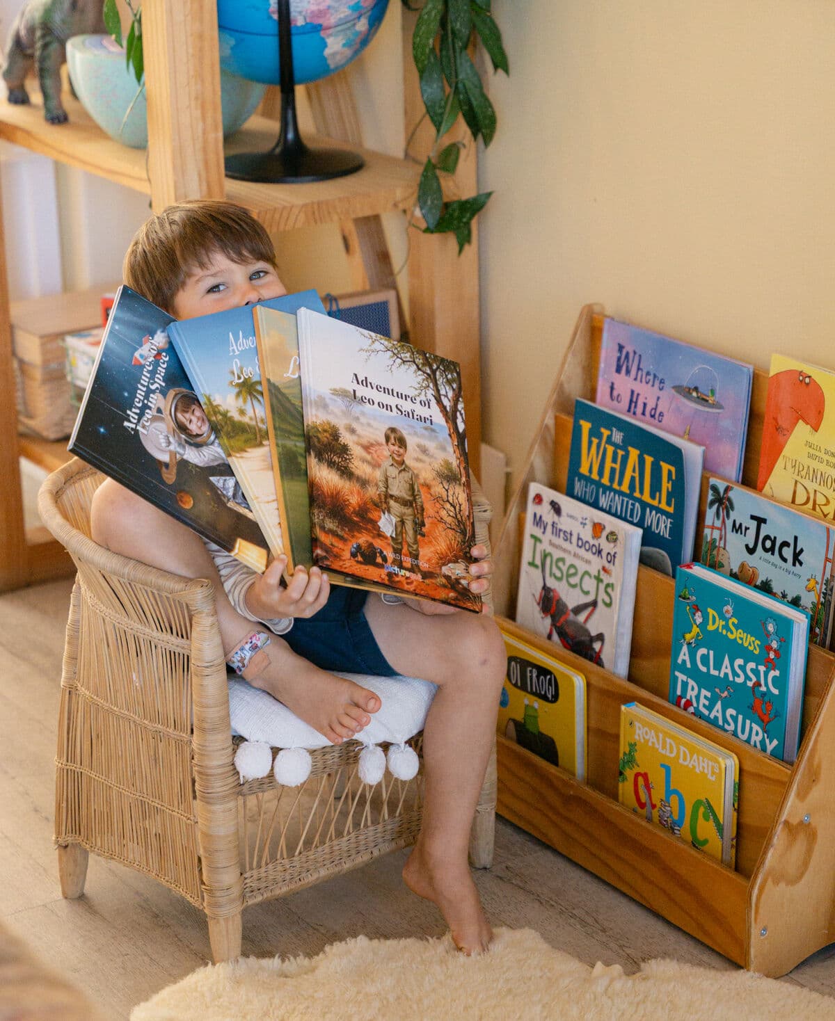 The boy peeking over all four of his personalised books, fanned out beside his bookshelf