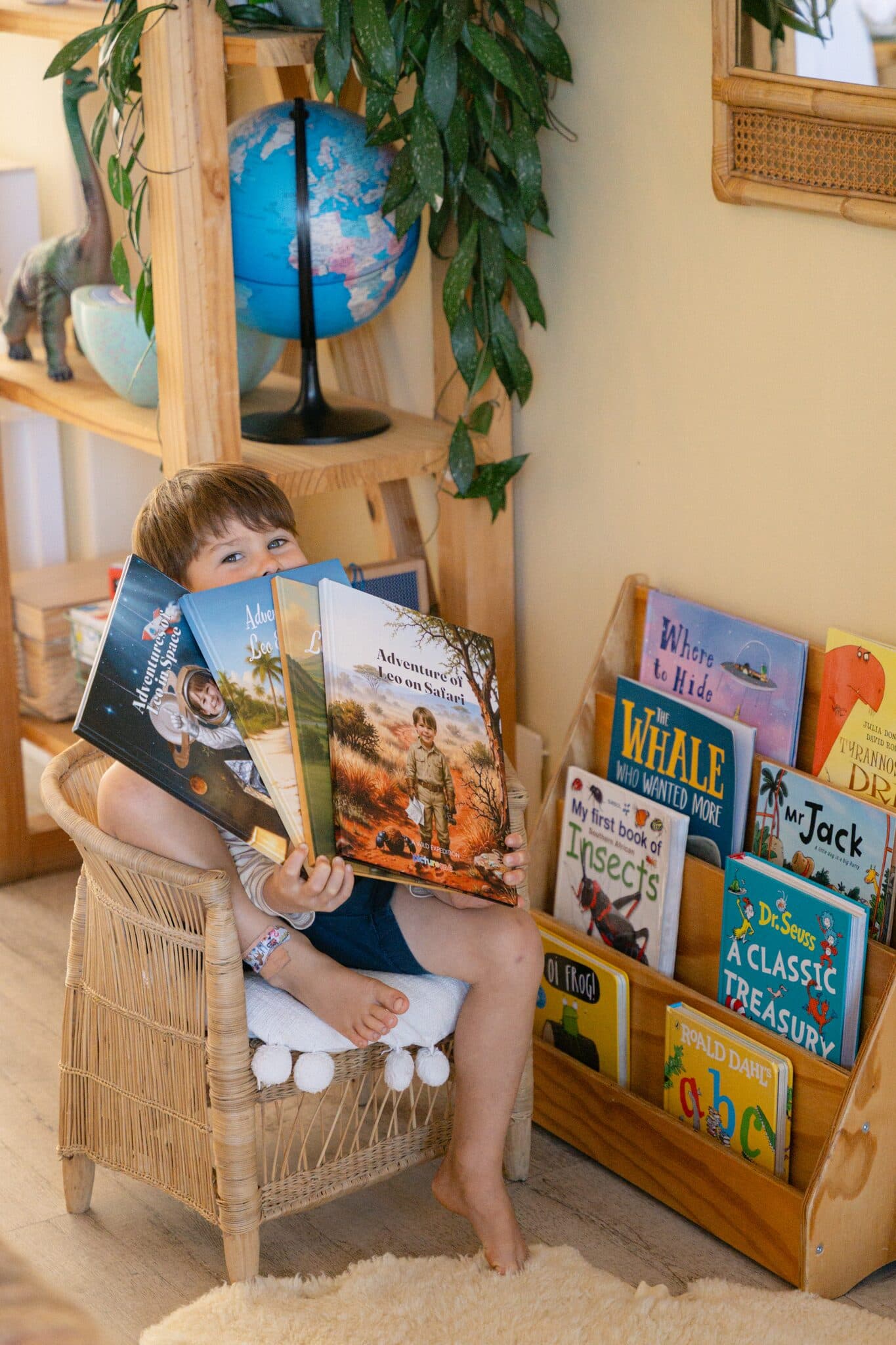 A boy in a reading chair holding four Picture Me hardcovers, beside a shelf of his other picture books